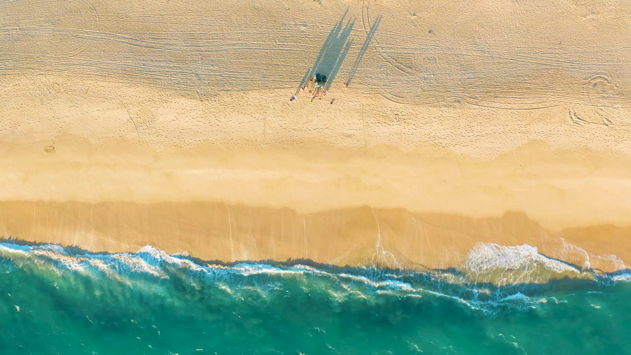 Top-down aerial frame over Diamante Cabo San Lucas with textured sand, a small shadowed mark near the upper beach, and curling turquoise surf forming scattered foam below.