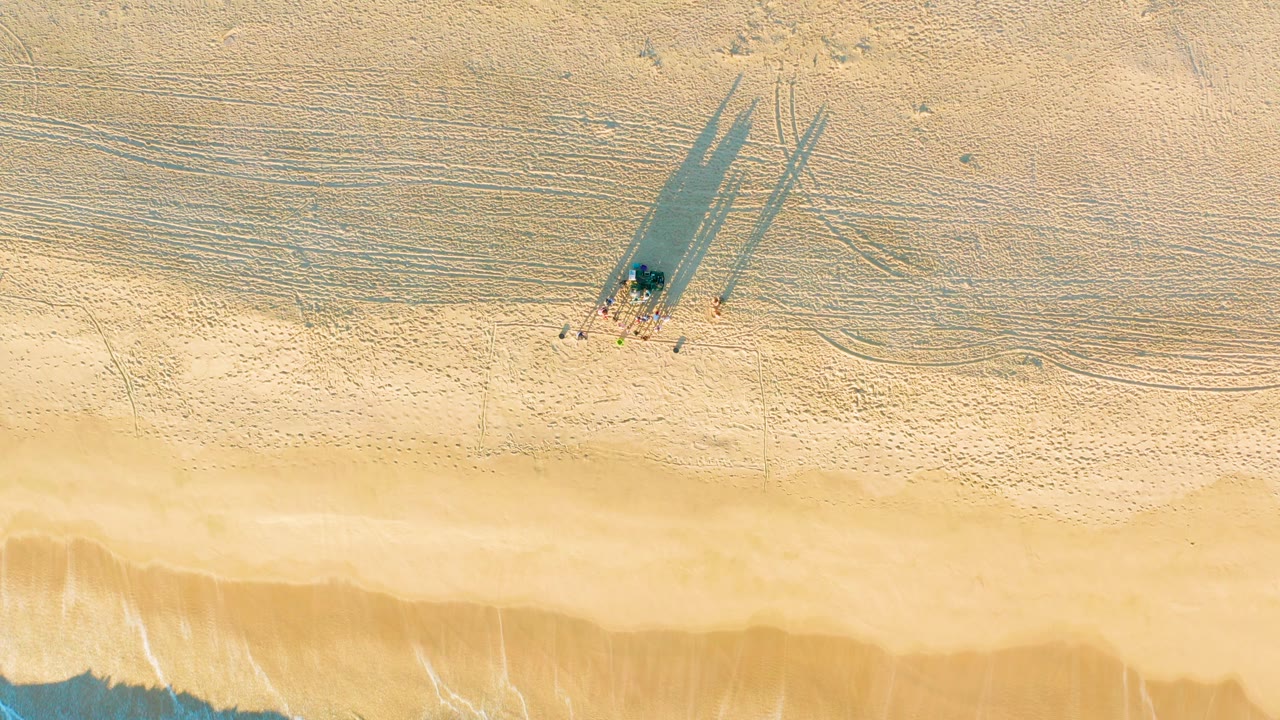 Top-down frame from Diamante Cabo San Lucas showing a small group walking across pale sand, their long shadows extending upward while the shoreline sits along the lower edge.