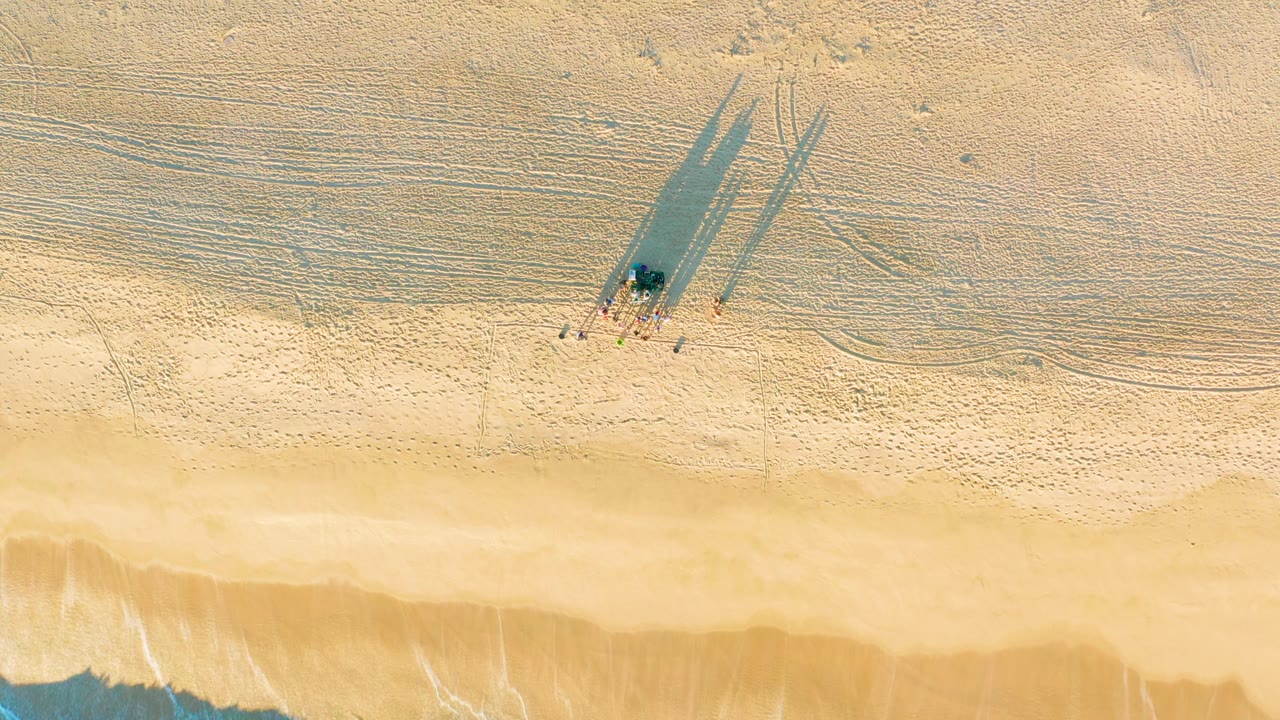 Aerial beach frame with walkers, long shadows, footprints, and faint surf at the bottom.