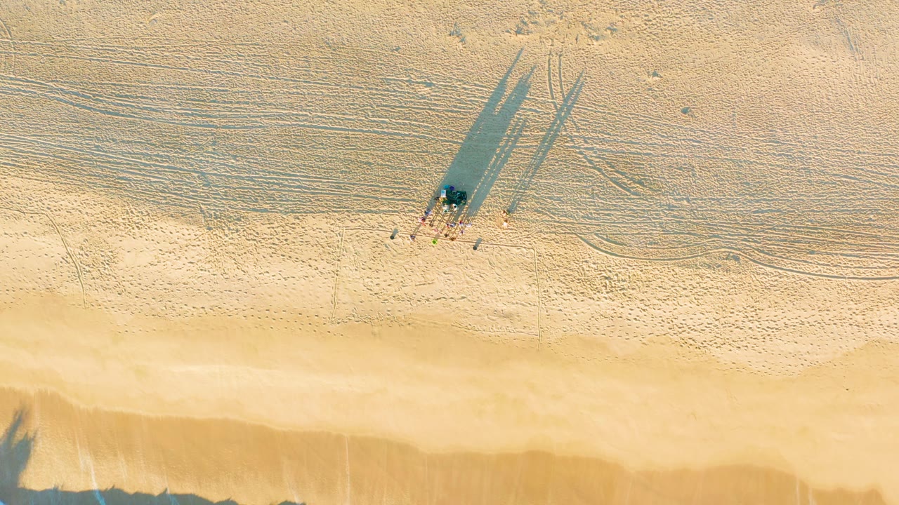 Aerial frame over Diamante Cabo San Lucas beach where the group remains near the upper sand and darker wet sand forms a broader strip before the waterline.