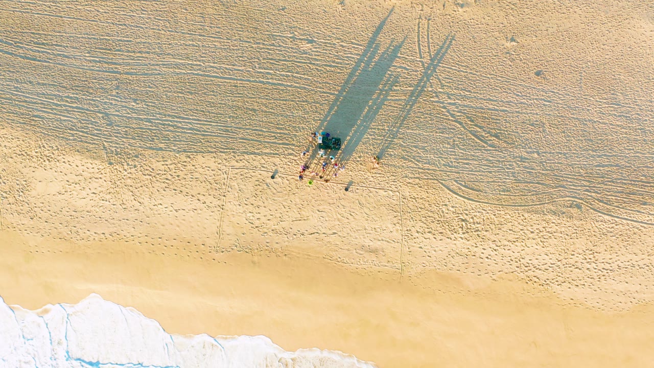 Drone view of beachgoers and long shadows beside retreating waves.