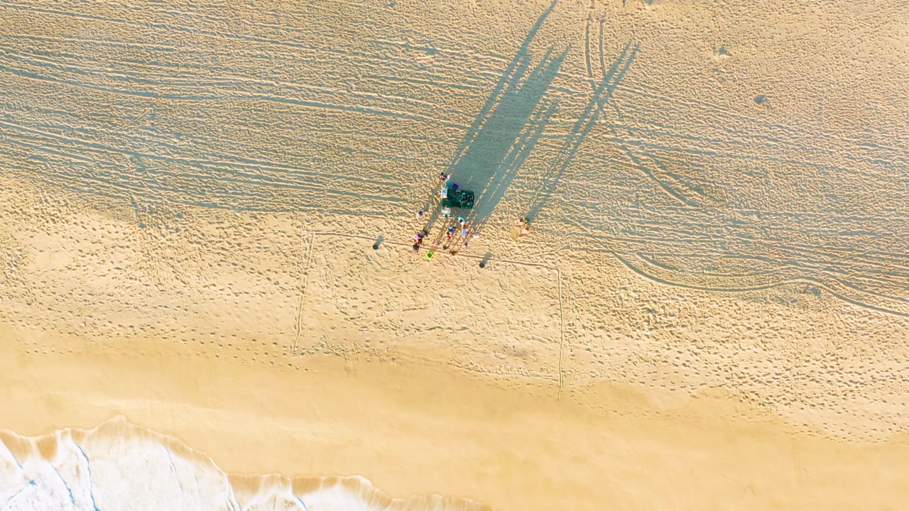 At 0:02, the overhead beach view shows the people as small figures beside a dark object or cluster on the sand, with elongated shadows and surf foam lower in the frame.