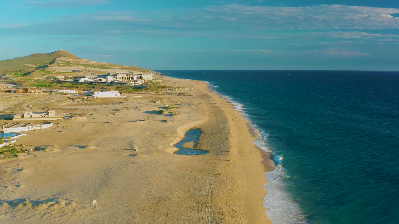 Aerial frame from Diamante Cabo San Lucas with the surf line running along the right edge, tide pools on the sand, distant buildings, a low hill, and no visible people.