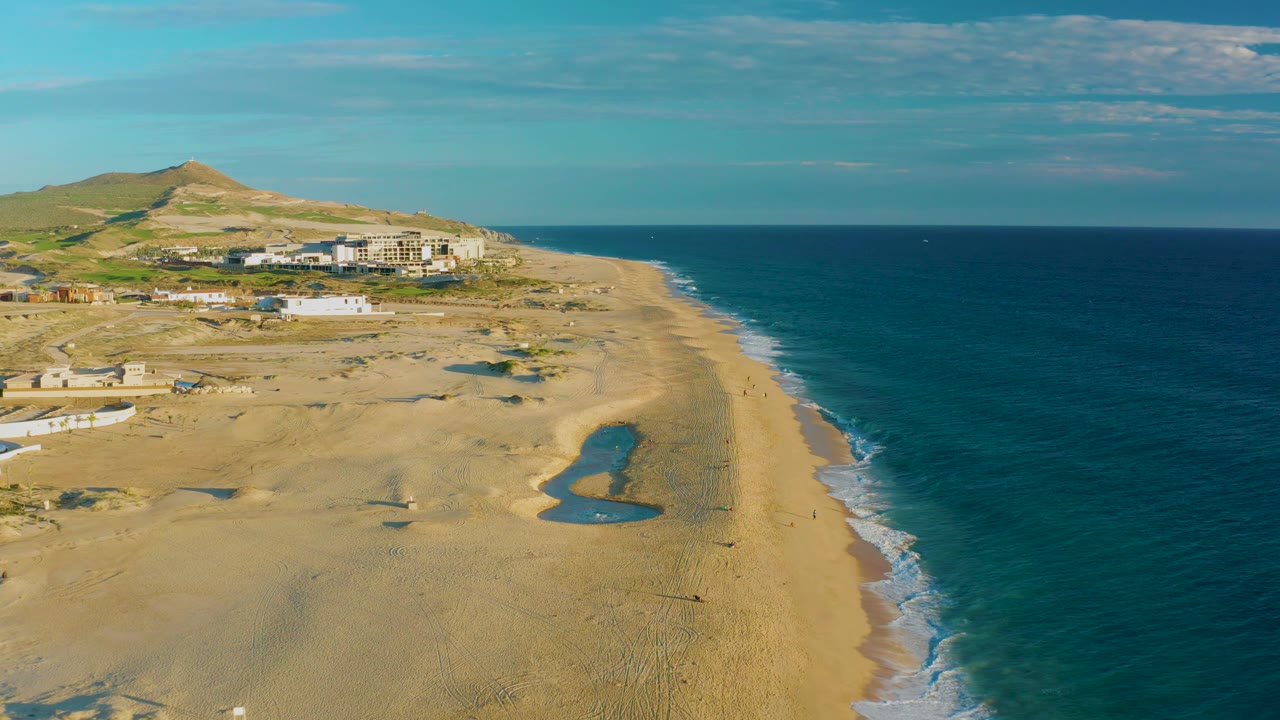 Sandy beach from above with turquoise ocean, tide pools, surf, buildings, and hill.
