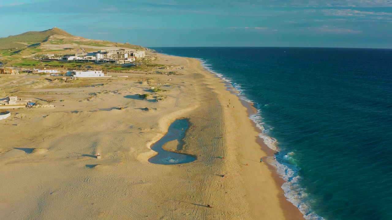 High drone view at Diamante Cabo San Lucas with the tide pools nearer the center, surf tracing the right edge of the beach, ridge buildings in back, and no visible people.