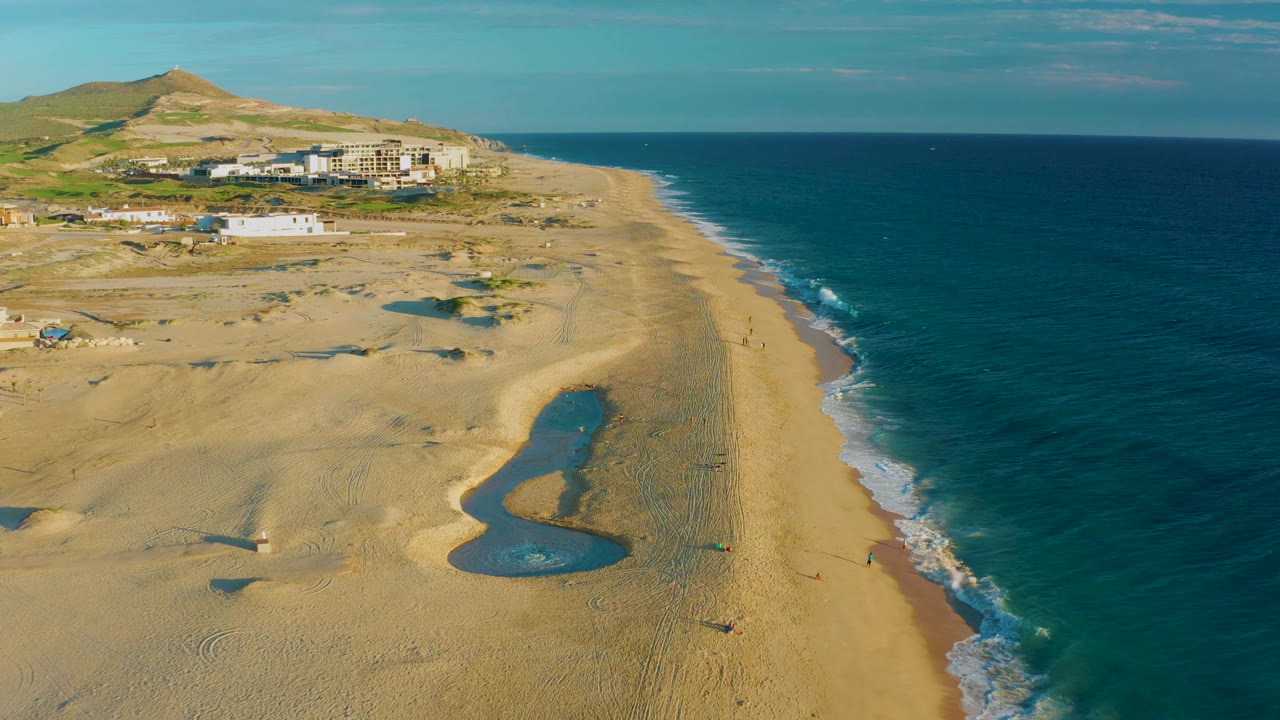 Aerial frame from Diamante Cabo San Lucas showing an elongated tide pool in the lower beach, small white waves on the shoreline, dune tracks, distant buildings, and no visible people.