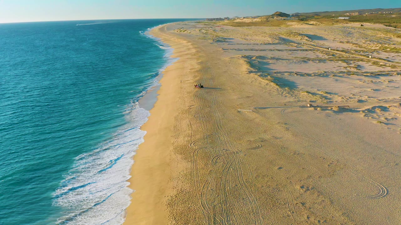Aerial frame over Diamante Cabo San Lucas showing turquoise water on the left, a pale sandy shoreline through the center, dune textures to the right, and no visible people.