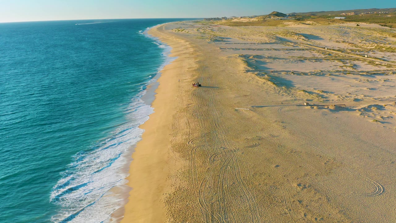 Aerial frame over Diamante Cabo San Lucas showing a broader sweep of beach tracks, turquoise surf to the left, dune ridges to the right, and no visible people.