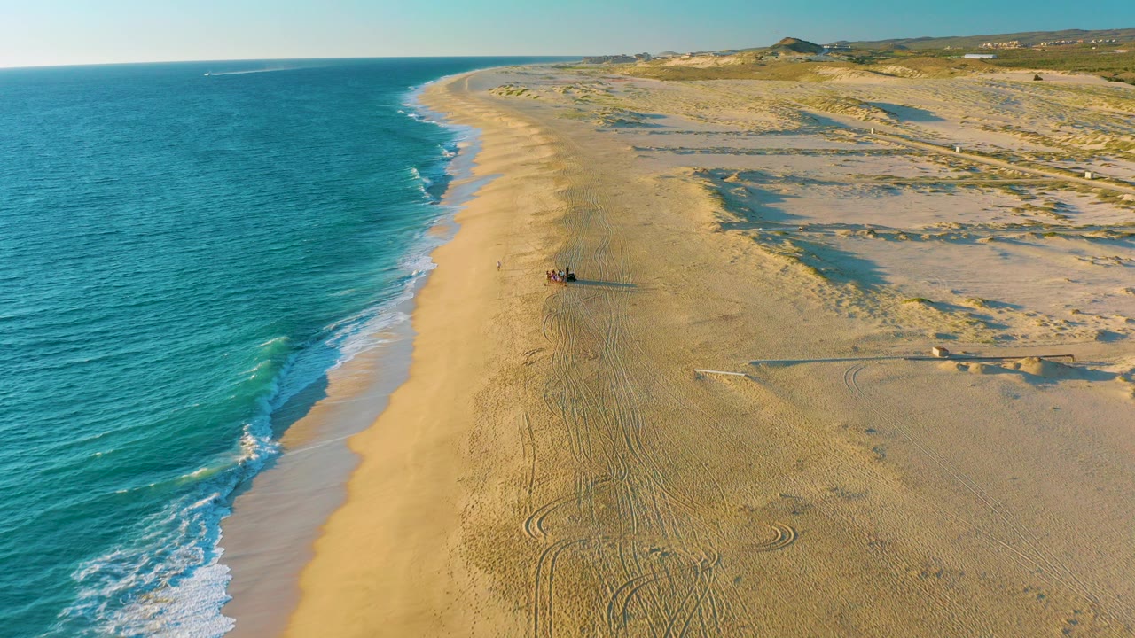 Aerial frame over Diamante Cabo San Lucas showing turquoise ocean on the left, a broad sandy beach, dune grass to the right, and small beachgoers near the center distance.