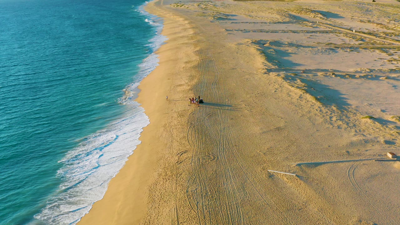 A high aerial frame over Diamante Cabo San Lucas shows a vehicle and small distant people on golden sand, blue ocean surf to the left, dunes to the right, and tire tracks trailing behind.