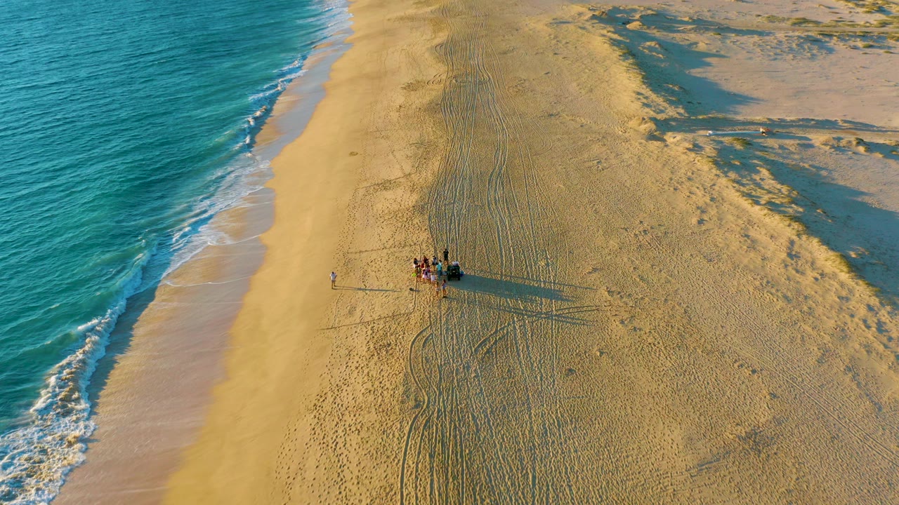 Aerial view at Diamante Cabo San Lucas showing turquoise surf at left, wide sand, a small group of people near a dark dune buggy, and long shadows stretching across the beach.