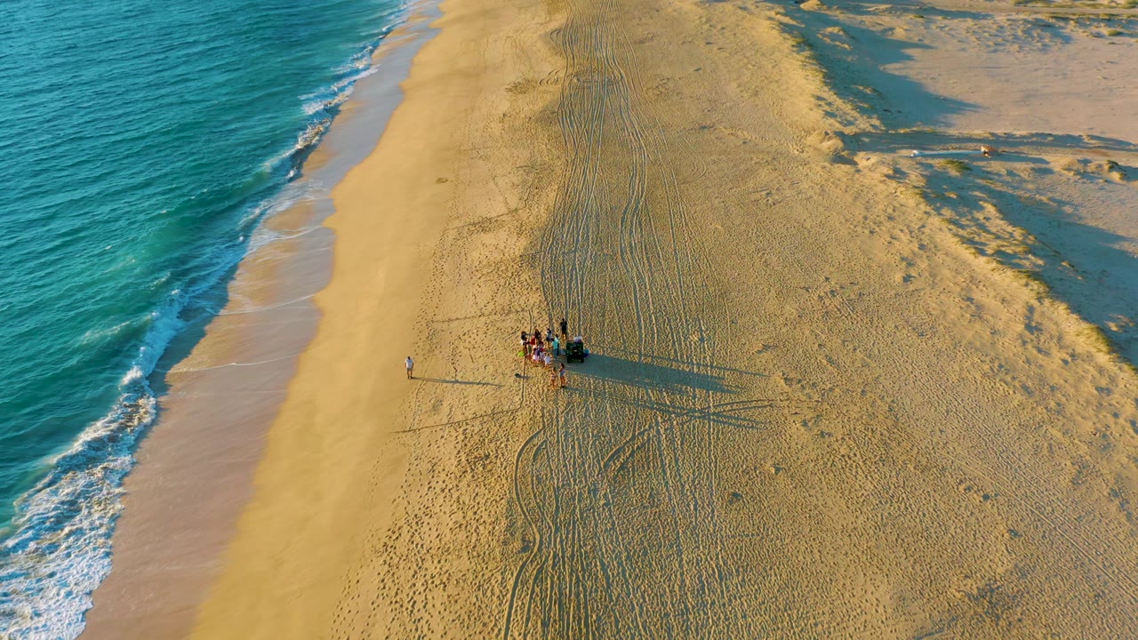 Aerial frame at Diamante Cabo San Lucas with the dune buggy and visible people slightly lower in the composition, wave wash at left, and textured beach sand and dunes to the right.