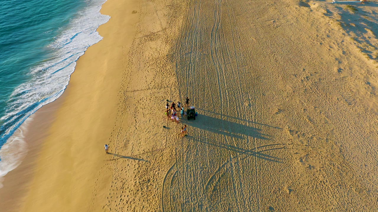 Overhead view of Diamante Cabo San Lucas beach at 0:00, with blue surf along the left edge, a small visible group near a dark vehicle, tire tracks in the sand, and long shadows stretching right.