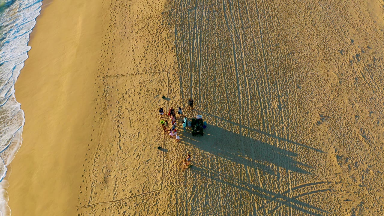 Overhead view of a small group of people on the sand at Diamante Cabo San Lucas, with surf along the left edge and long shadows reaching across the beach.