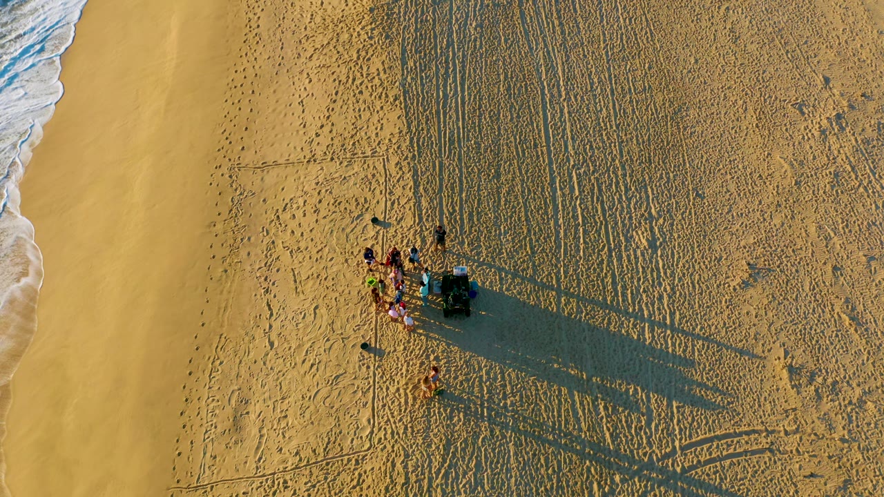 Drone view at Diamante Cabo San Lucas with the people and gear slightly right of center, white surf at the left, and parallel sand textures filling the upper beach.