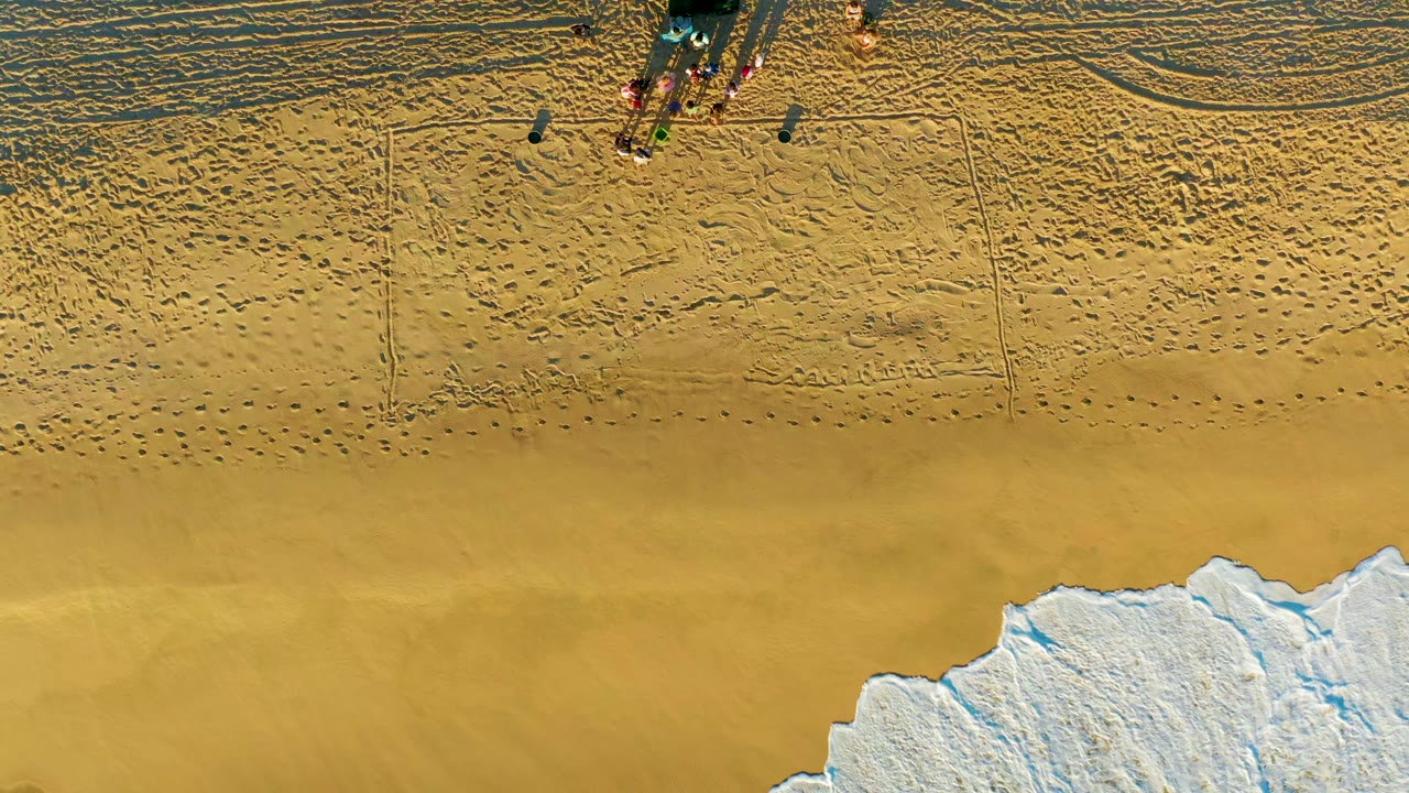 Overhead view at Diamante Cabo San Lucas showing a dark vehicle and small group on patterned sand while foamy surf reaches the lower edge of the frame.