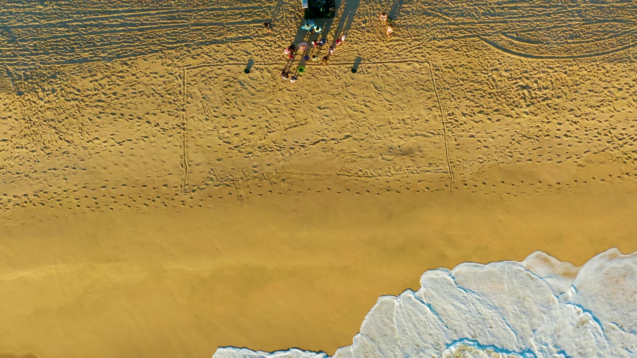 Aerial frame at Diamante Cabo San Lucas showing the group and vehicle on textured sand as surf foam shifts farther across the lower right corner.