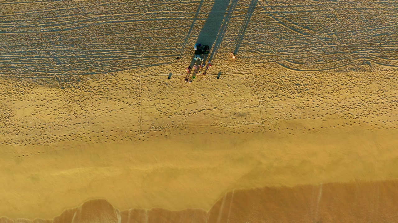 Overhead view of a sandy beach in Diamante Cabo San Lucas with a small group near the upper frame, long shadows across the sand, and darker wet shoreline below.