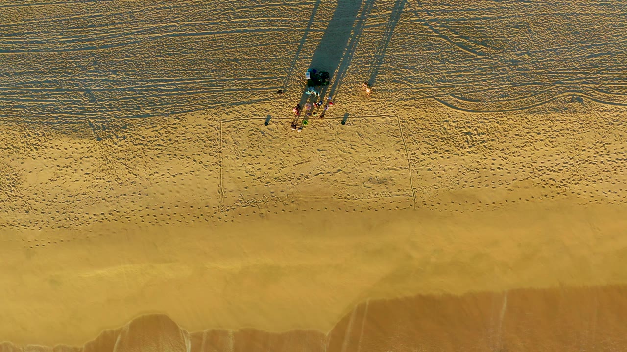 Aerial beach frame with people casting shadows and waves lower in frame.