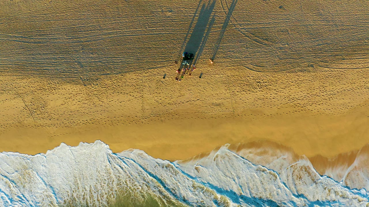 Aerial beach view with foam line, people, structure, and sand patterns.