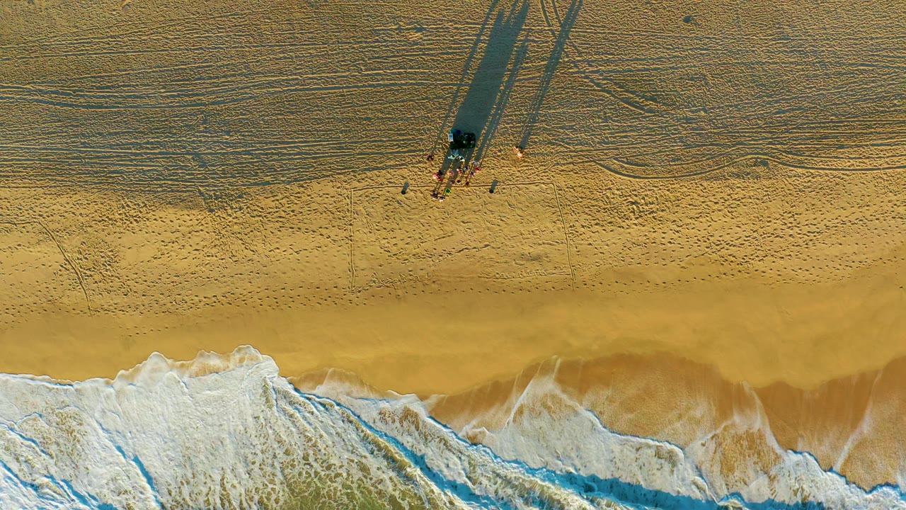 Top-down view of the Diamante Cabo San Lucas shoreline with white water along the bottom, open sand, and a small group beside a structure throwing long shadows across the beach.
