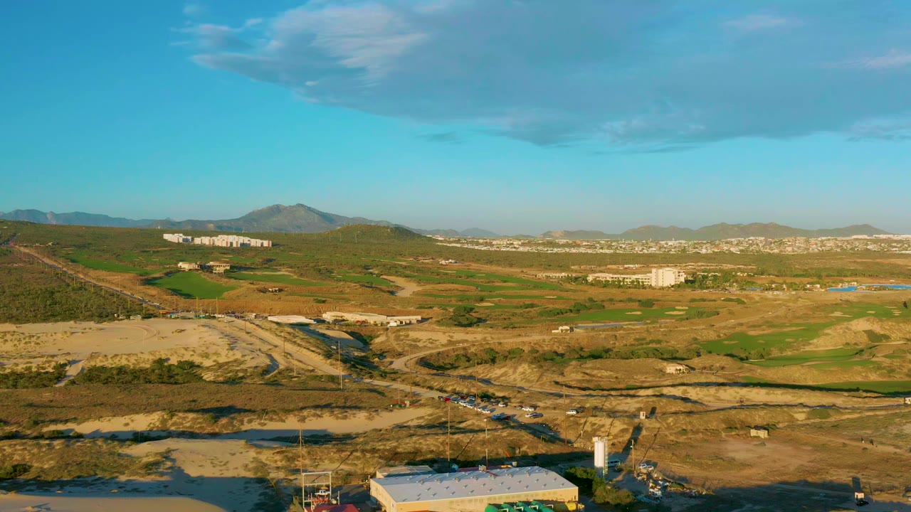Wide aerial frame over Diamante Cabo San Lucas, Baja California Sur, showing green and dry fields, dirt roads, scattered buildings, distant mountains, blue sky, and no visible people.