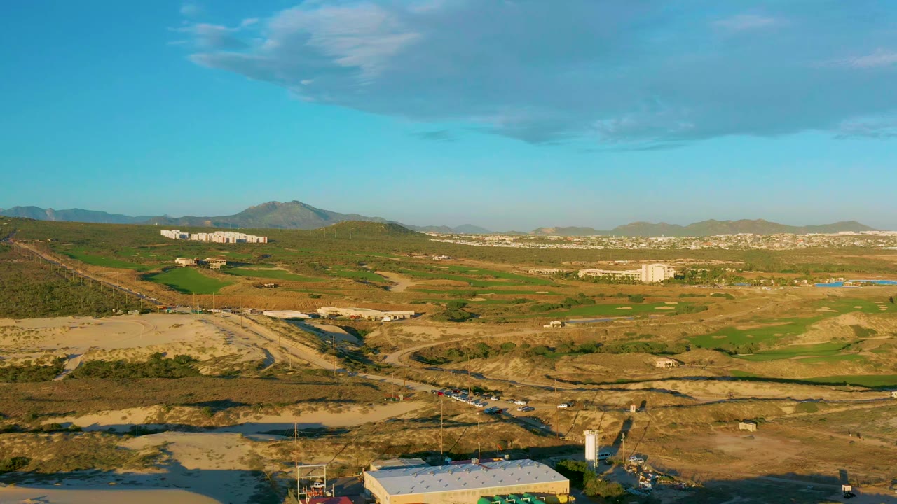 Aerial landscape with open fields, road cuts, low buildings, and mountains below a clear blue sky.
