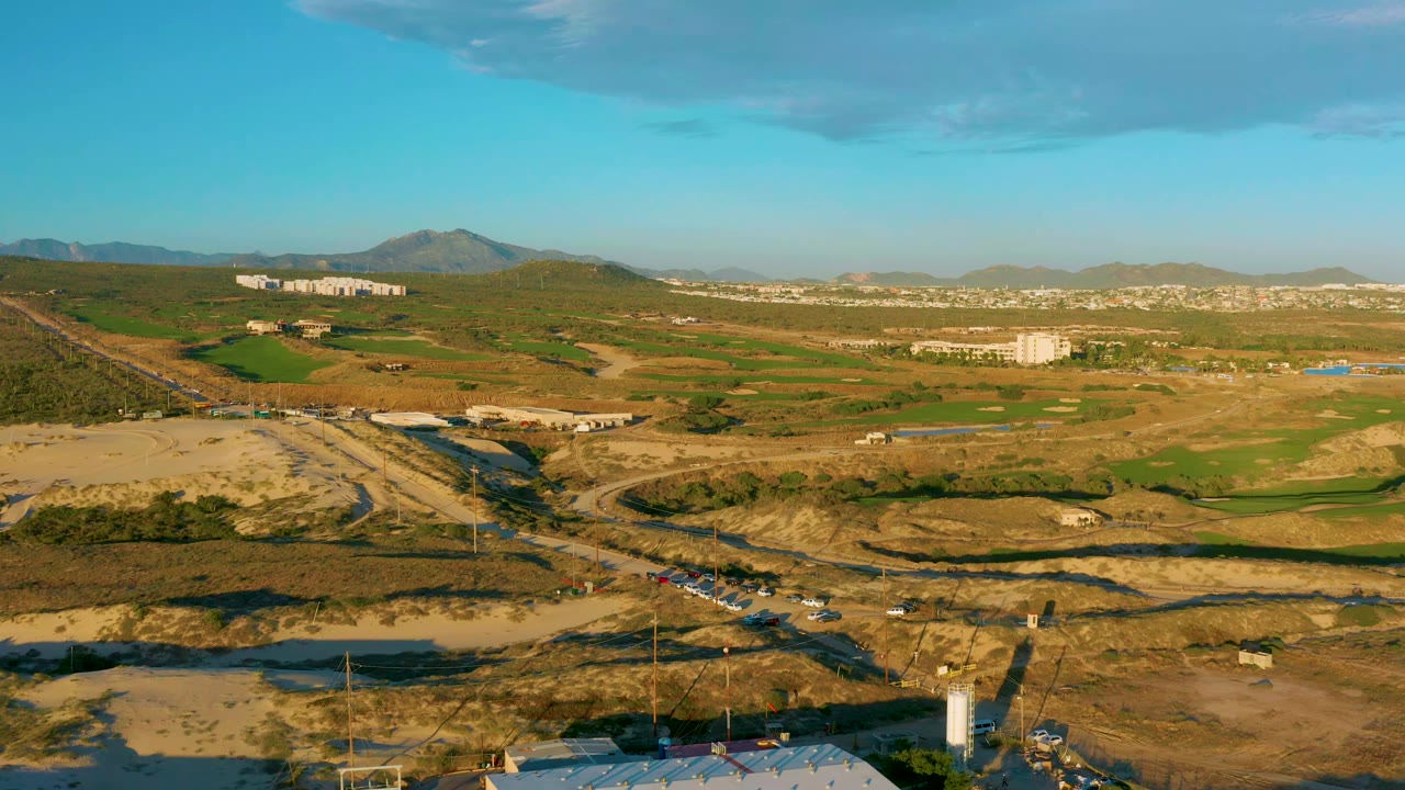 Aerial frame in Diamante Cabo San Lucas showing graded dirt roads, patchwork green fields, low buildings, distant residential-looking blocks, mountains, and no visible people.