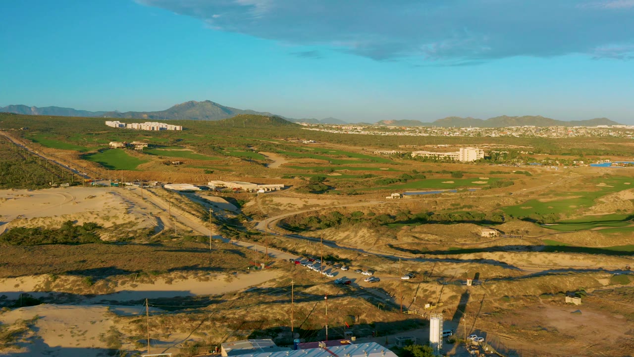 High drone view over Diamante Cabo San Lucas with a dirt site and road in the foreground, small vehicles near the road, fields beyond, mountains in back, and no visible people.