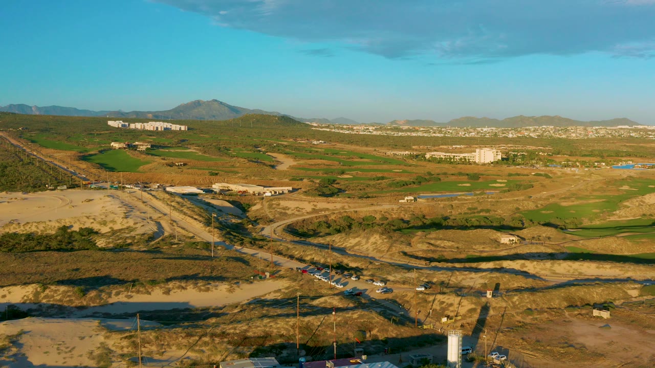 Aerial construction landscape with dirt lots, green fields, road curves, and distant buildings.