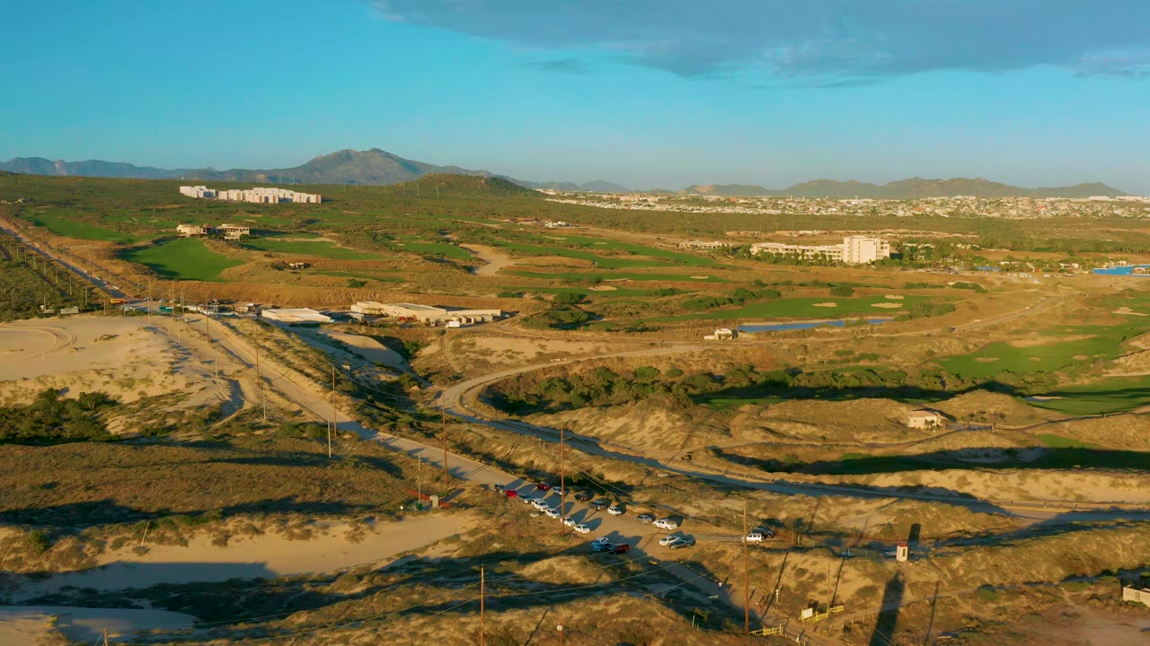 Drone view in Diamante Cabo San Lucas with vehicles clustered near the lower road, a curved road through dry ground, green midground patches, distant white buildings, mountains, and no visible people.