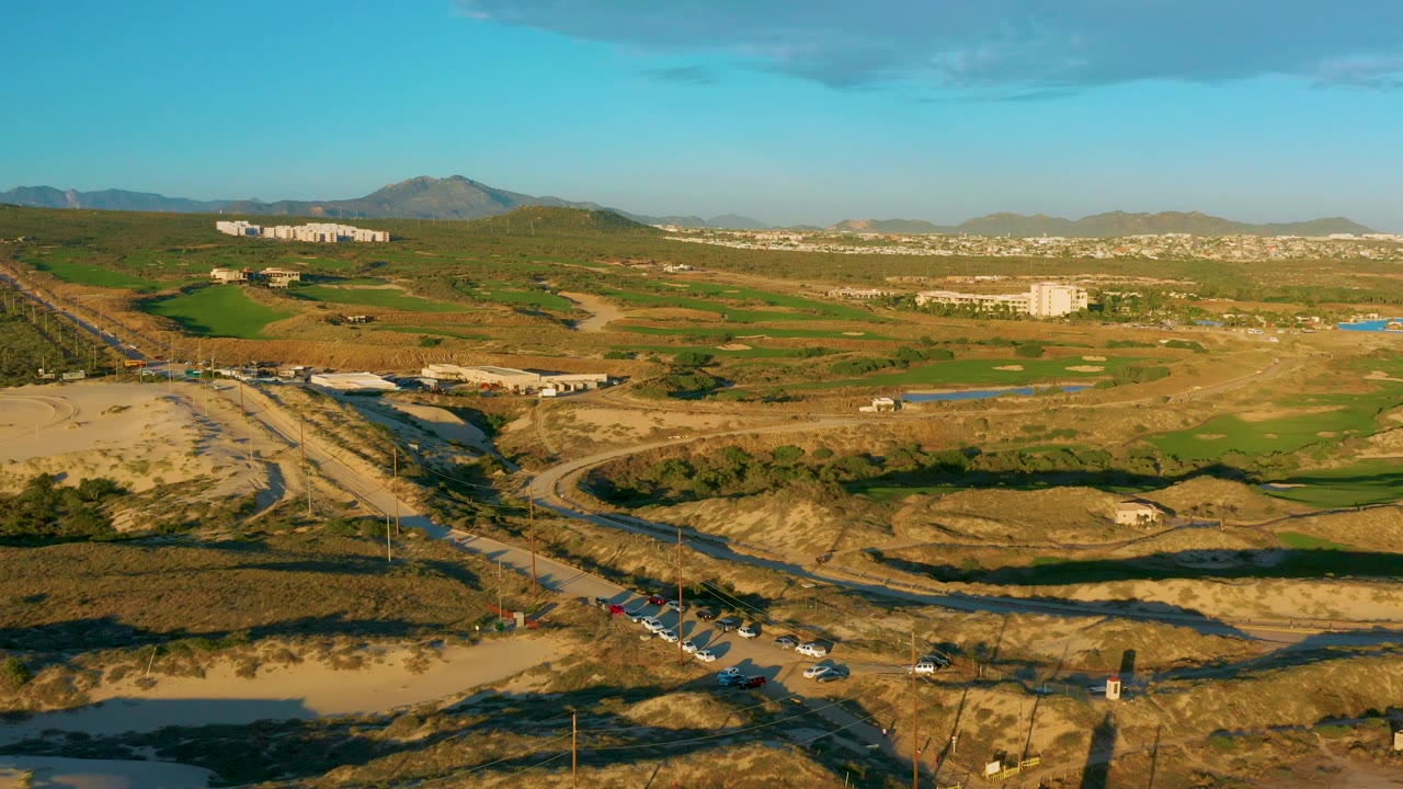 Wide aerial frame over Diamante Cabo San Lucas with cars along the lower road, dry lots and green land across the midground, white structures in the distance, hills, mountains, and no visible people.
