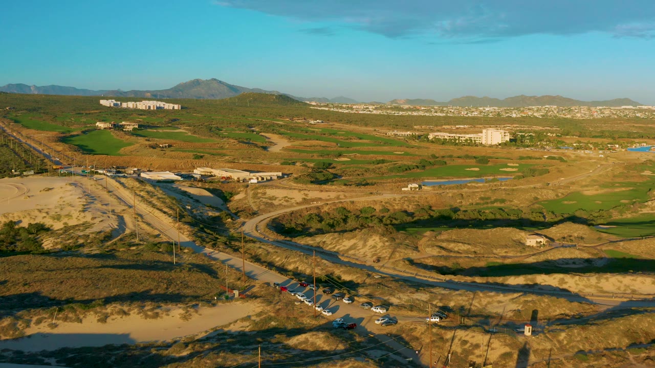 Aerial view of Diamante Cabo San Lucas with vehicles moving along the lower road, long shadows across dry ground, green patches in the midground, low buildings, mountains, and no visible people.