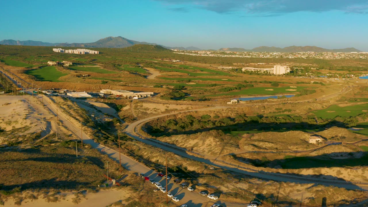 Aerial frame over Diamante Cabo San Lucas showing a curved road, dry fields, green patches, distant buildings, low hills, blue sky, and no visible people.