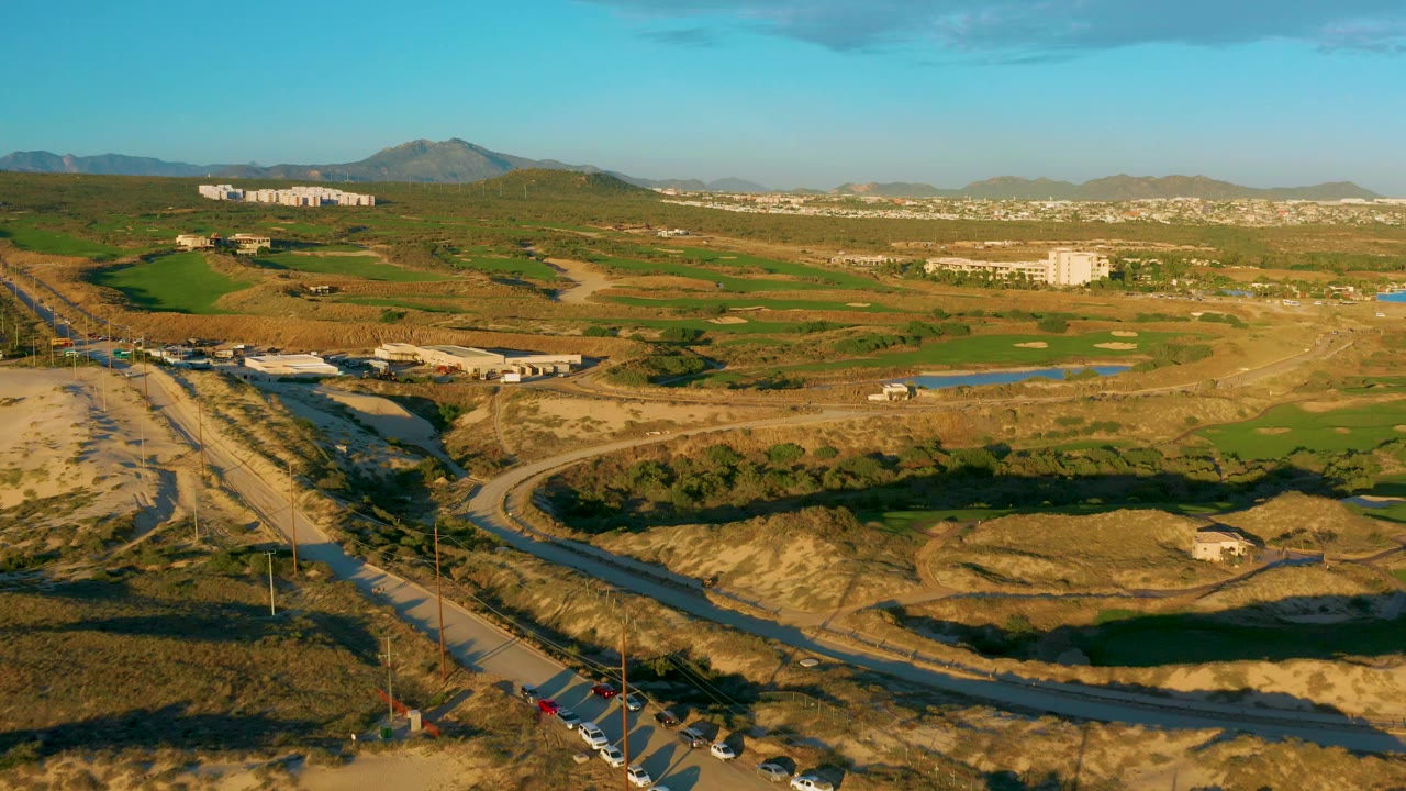 Drone-height view over Diamante Cabo San Lucas with a road and bridge area in the foreground, tan ground, green fields, small water features, distant buildings, hills, and no visible people.