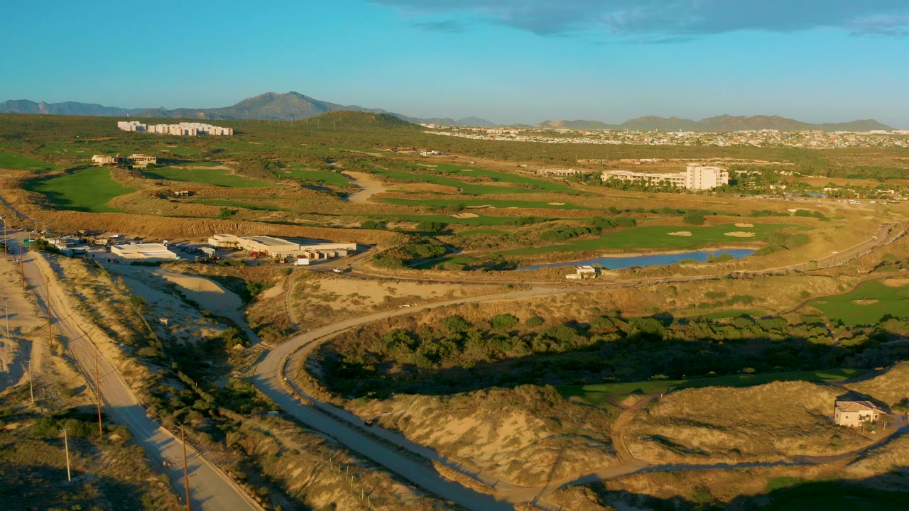 Aerial view over Diamante Cabo San Lucas with a winding road in the foreground, green fields and ponds in the middle distance, distant hills, and no visible people.