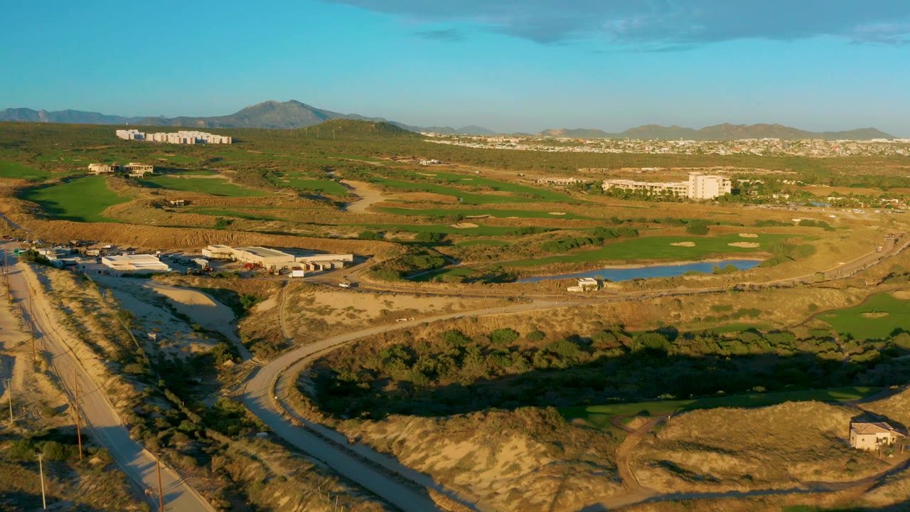 Wide aerial frame over Diamante Cabo San Lucas with a foreground road, patchwork green fields, ponds near the center right, distant buildings, low mountains, and no visible people.