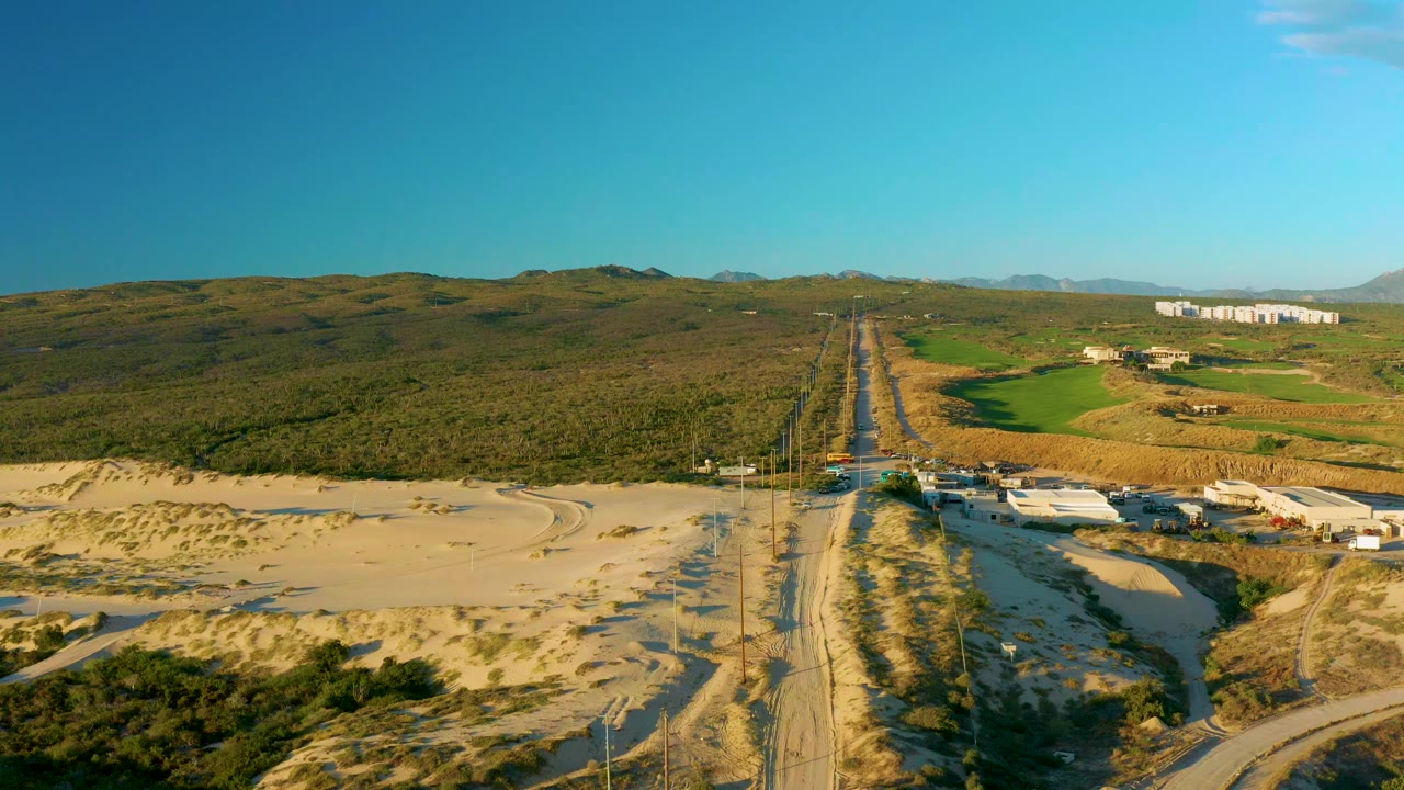 Opening aerial frame of a straight highway in Diamante Cabo San Lucas, Baja California Sur, with sandy ground, green fields, distant hills, clear blue sky, and no visible people.