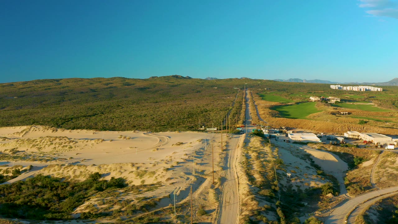 Aerial frame of the Diamante Cabo San Lucas highway with utility poles lining the road, dry sand and scrub in the foreground, fields and low structures to the side, and no visible people.