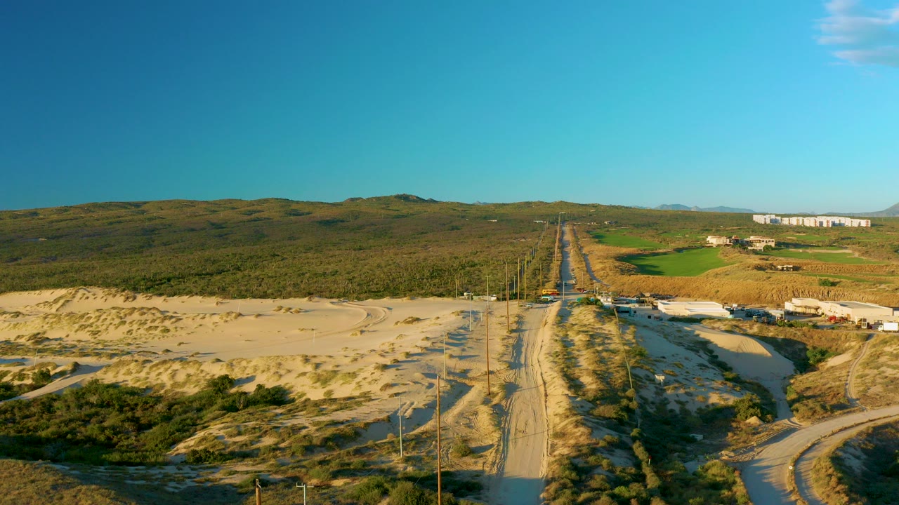 Aerial view near Diamante Cabo San Lucas showing a sandy road centered between dunes and utility poles, with low arid hills in the distance and no visible people.