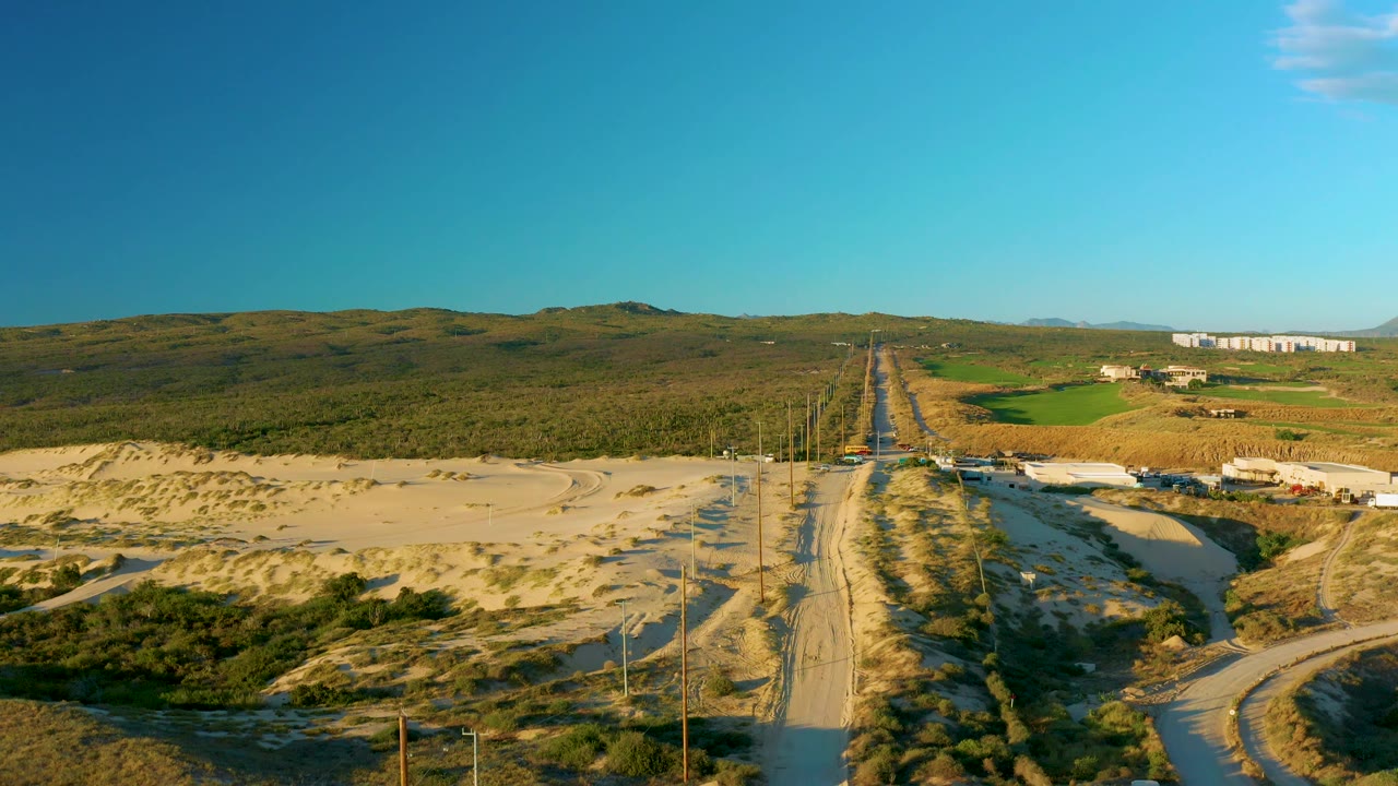 Aerial view near Diamante Cabo San Lucas showing the sandy road, dune vegetation, a few small buildings on the right side, distant hills, and no visible people.