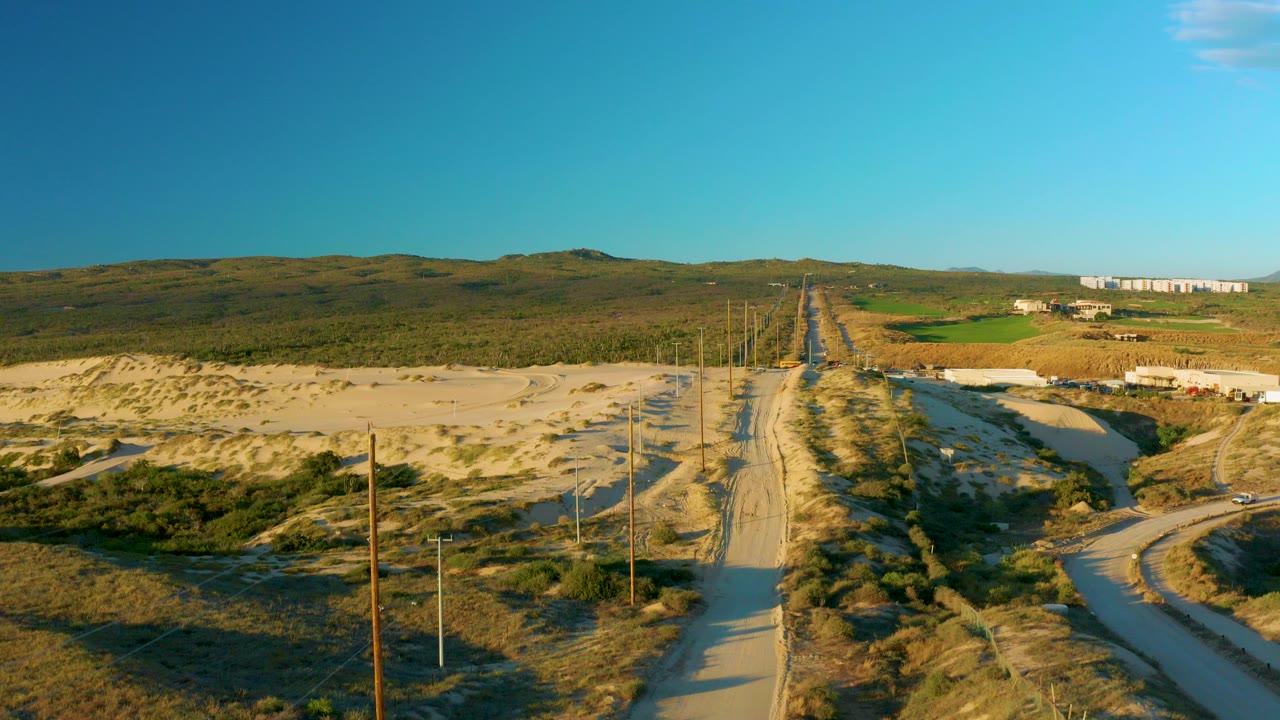 Aerial view in Diamante Cabo San Lucas, Baja California Sur, showing a sandy road running between scrub-covered dunes and utility poles with no visible people in the frame.