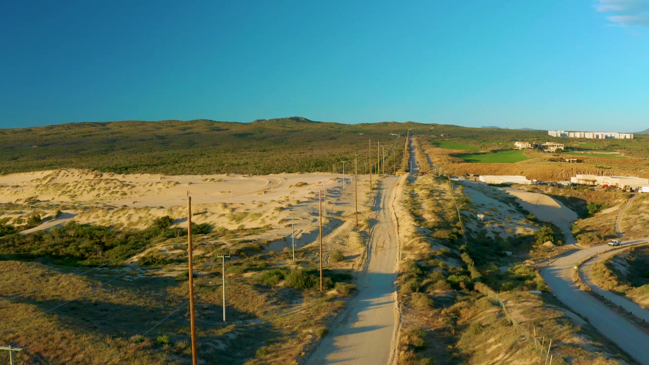 Aerial road view in Diamante Cabo San Lucas, Baja California Sur, with utility poles receding beside a sandy track, dry vegetation on both sides, and no visible people in the frame.