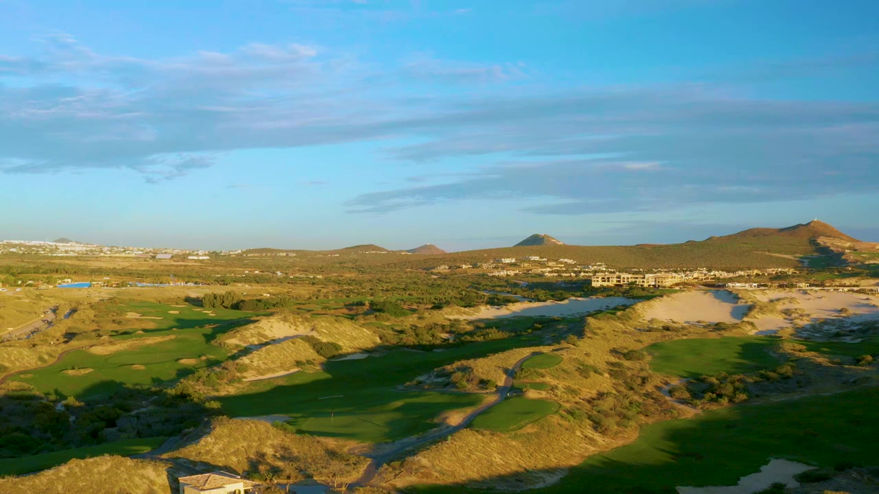 Opening aerial frame over Diamante Cabo San Lucas shows green golf course fairways across dry rolling hills, distant low buildings, broad blue sky, and no visible people.