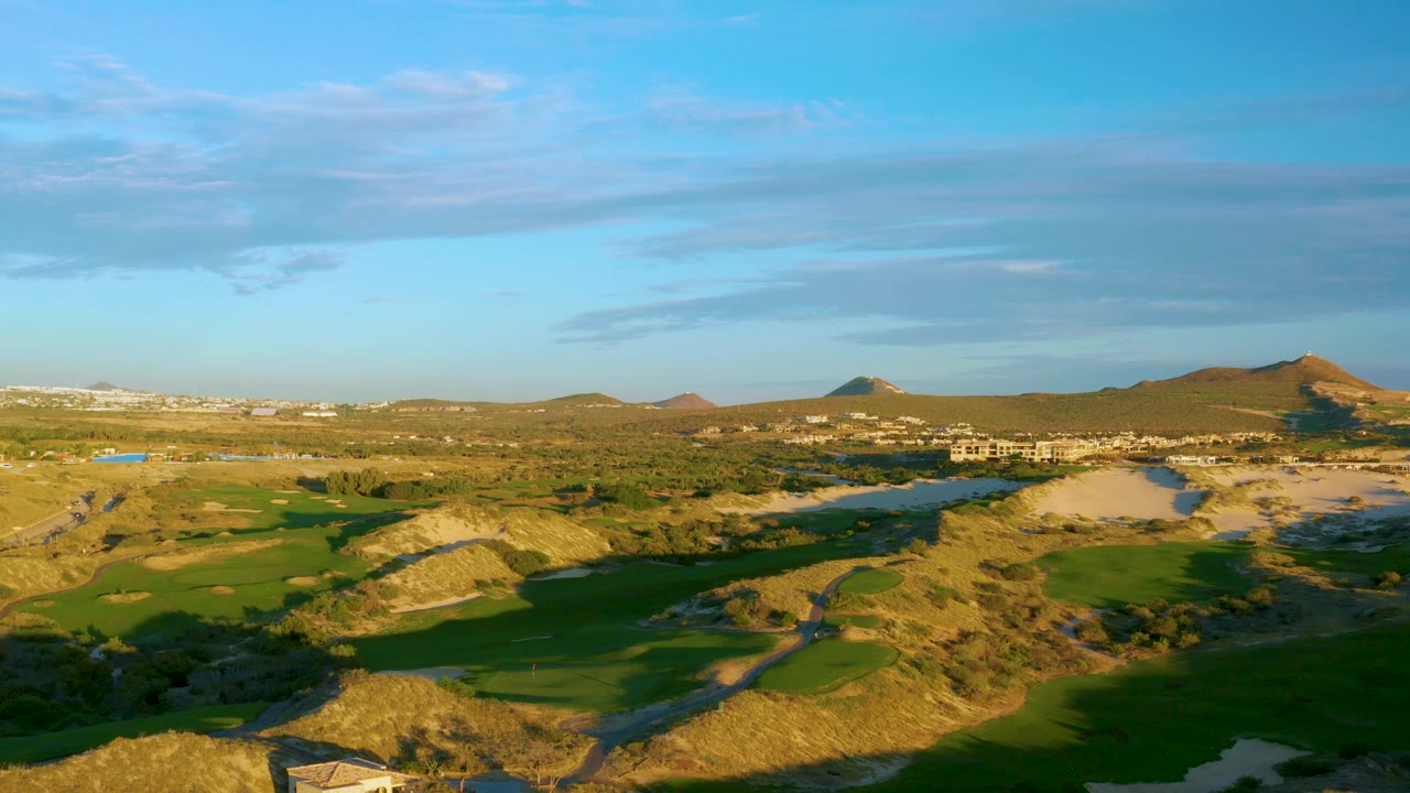 At 0:02, the aerial view shows sunlit tan hills and green golf course sections in Diamante Cabo San Lucas, with sandy patches, distant buildings, and no visible people.