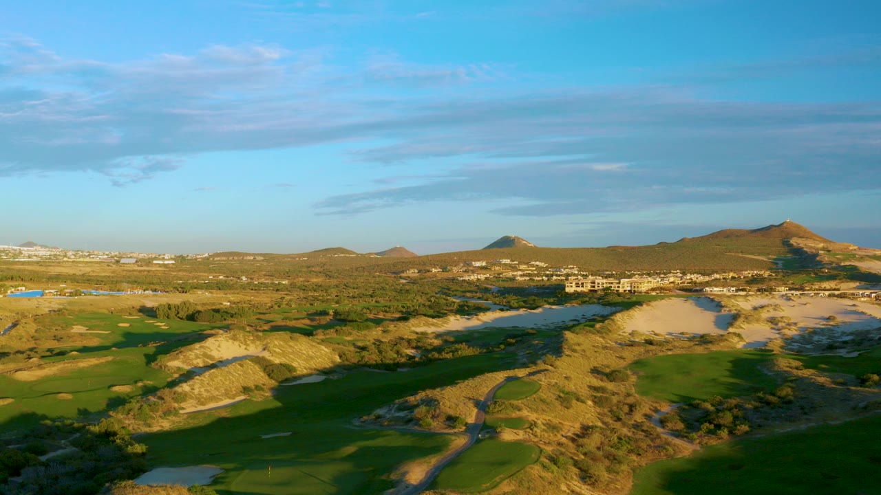 Opening frame over a Diamante Cabo San Lucas golf course with green fairways, sandy traps, scrub hills, distant buildings, low mountains, and no visible people.