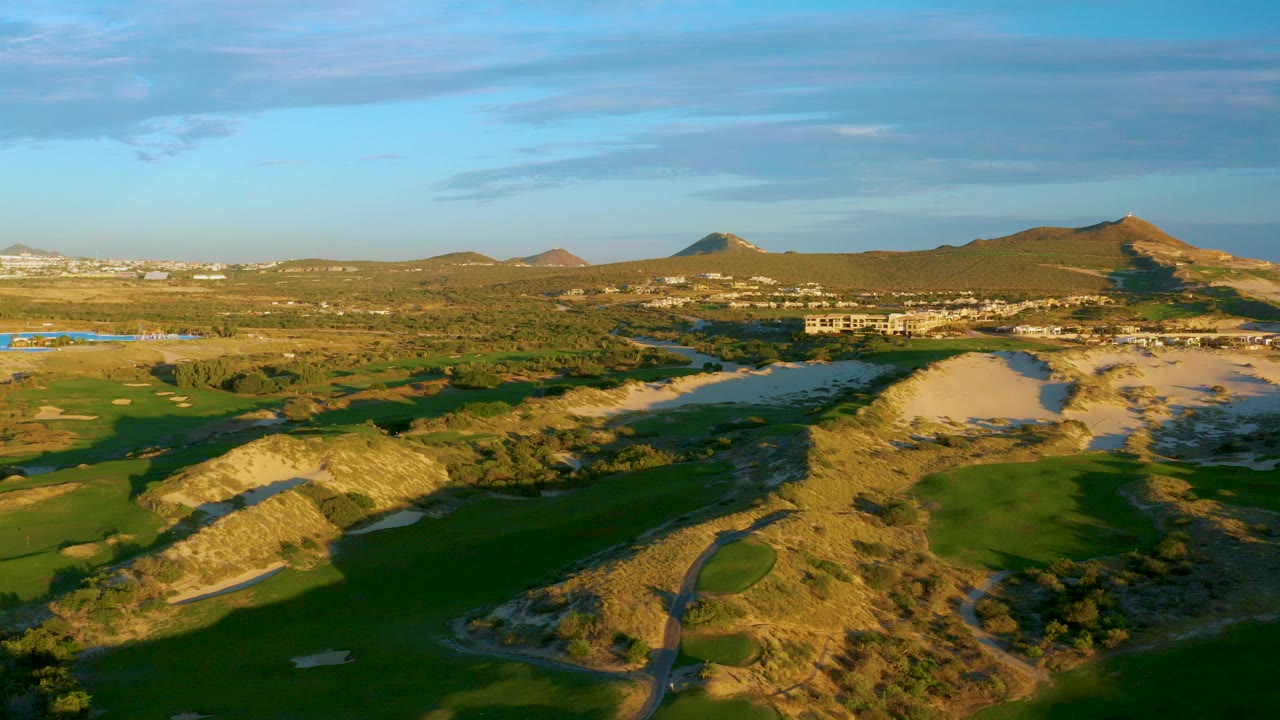 Aerial frame from Diamante Cabo San Lucas showing green golf fairways, pale sandy dunes, scattered low buildings, distant hills, blue sky, and no visible people.