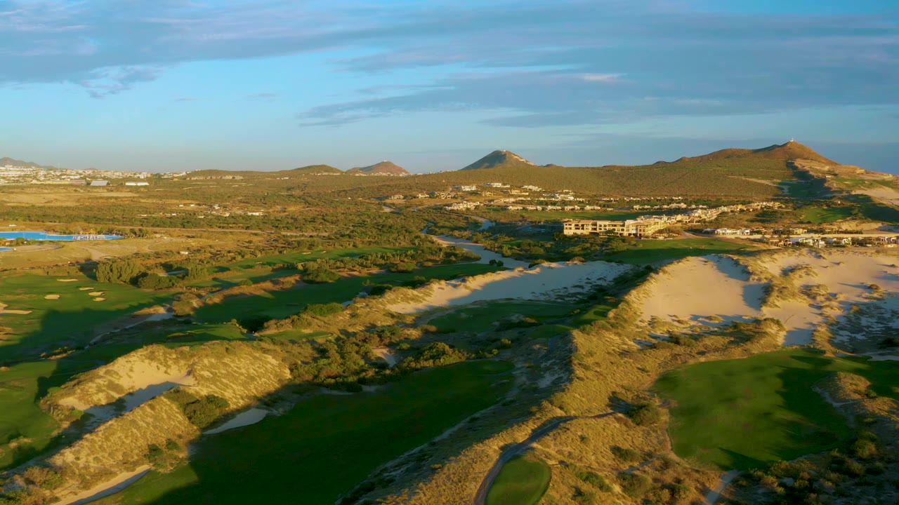 One second into the clip, the Diamante Cabo San Lucas aerial holds on fairways divided by sandy dunes, with water in the midground, hills beyond, and no visible people.