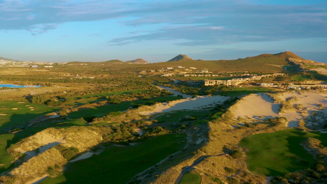 Midway through the clip, Diamante Cabo San Lucas appears in warm light with dark fairway shadows, bright sand dunes, water, low buildings, hills, and no visible people.