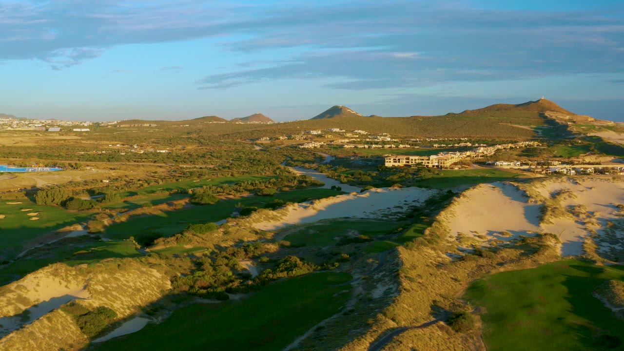 Opening aerial frame over Diamante Cabo San Lucas with green fields, sandy dunes, a winding river corridor, distant buildings, low hills, warm sunlight, and no visible people.